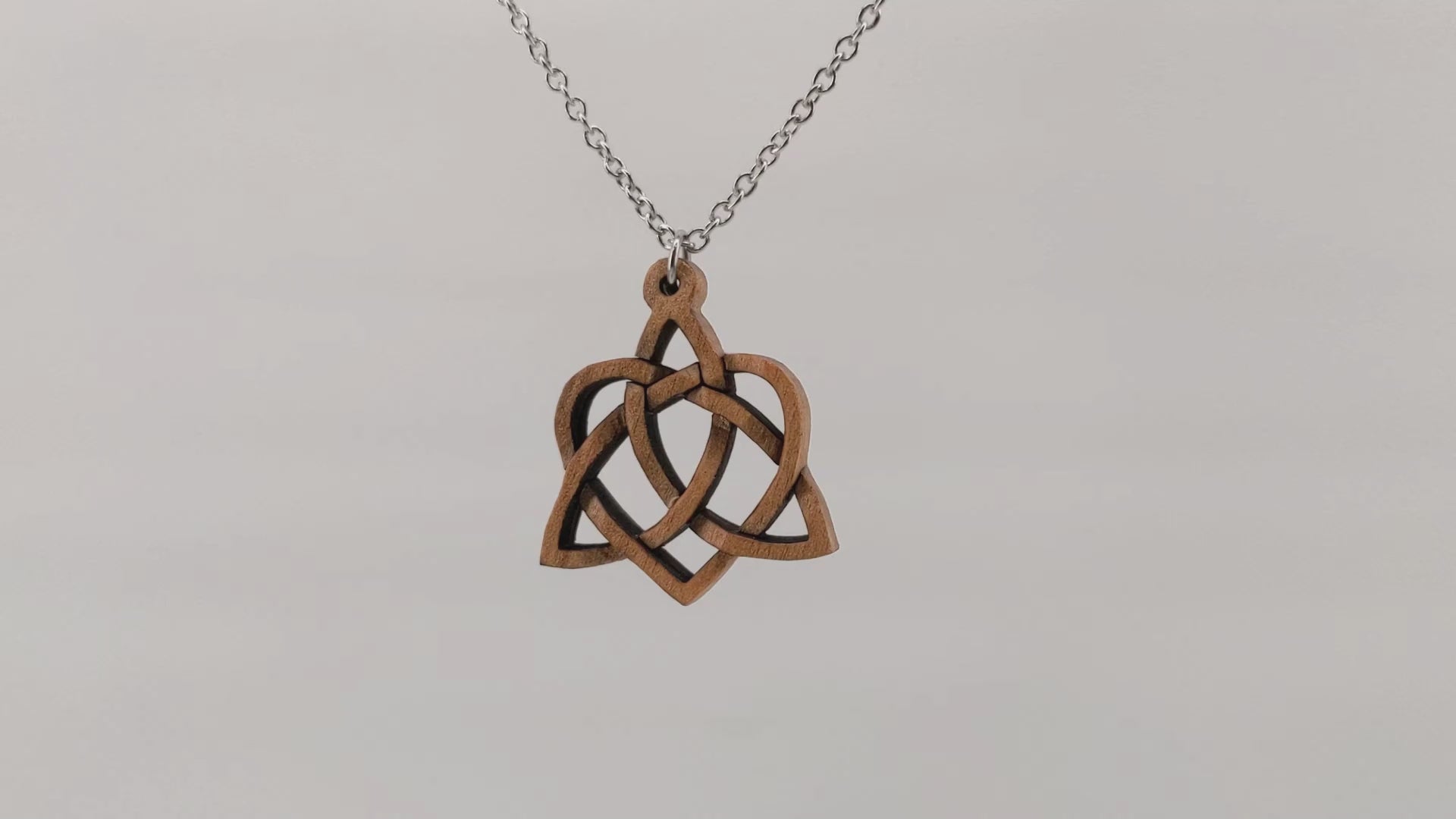 Wooden necklace pendant created in the style of a Celtic weave pattern that is a trinity and heart combined. The symbol represents adoption. Hanging and rotating from a silver stainless steel chain against a white background.
