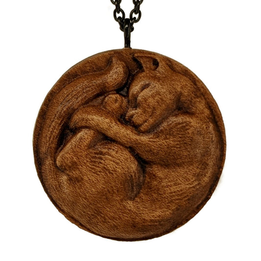 Wooden necklace pendant carved into the shape of a cat curled into a round ball. Hanging from a black stainless steel chain against a white background.