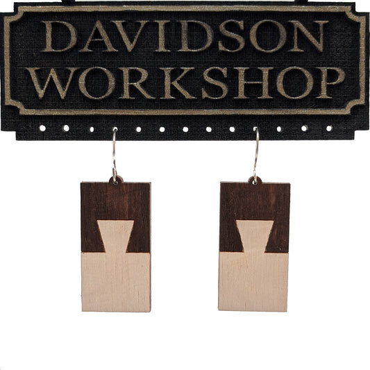 Pair of wooden earrings with silver stainless steel hooks. They are rectangular with a two toned dark brown and pale dovetail joinery design. Made from birch wood hanging from a model Davidson Workshop sign against a white background. (Close up View)
