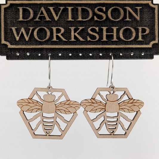 Pair of wooden earrings with silver stainless steel hooks. They are natural finished honeybees with its arms and wings spread on a honeycomb. Made from birch wood hanging from a model Davidson Workshop sign against a white background (Close up View)