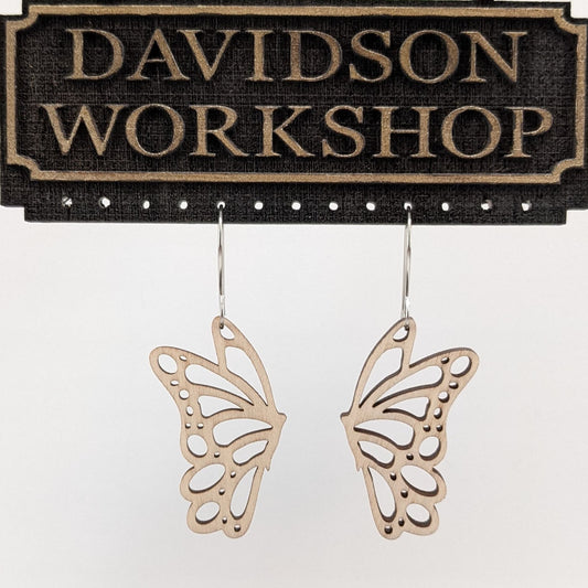 Pair of wooden earrings with silver stainless steel hooks. They are pale natural finished cutouts of side profile butterflies. Made from birch wood hanging from a model Davidson Workshop sign against a white background. (Close up View)