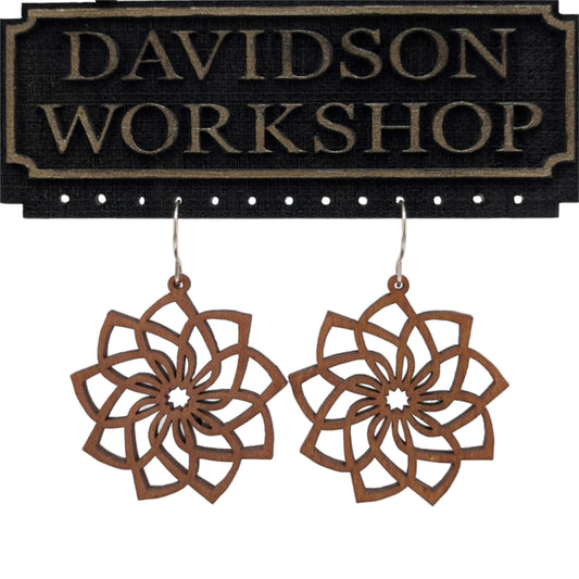 Pair of wooden earrings with silver stainless steel hooks. They are brown cutouts of flowers with spiraling, overlapping petals. Made from birch wood hanging from a model Davidson Workshop sign against a white background. (Close up View)