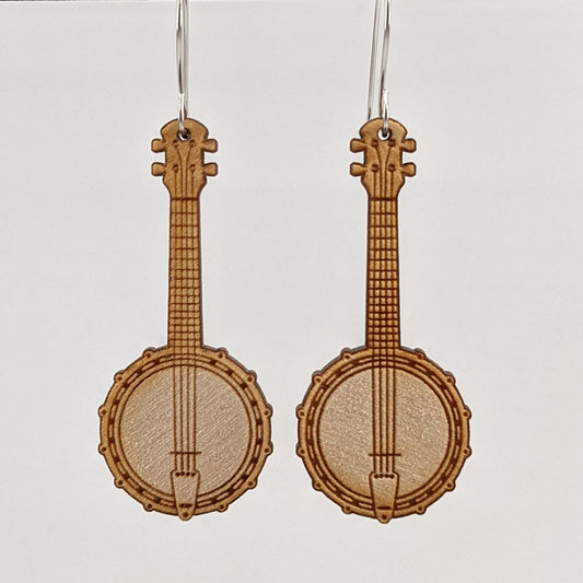 Pair of wooden earrings with silver stainless steel hooks. They are pale natural finished miniature 4 string banjos with two pickups. Made from birch wood hanging from a model Davidson Workshop sign against a white background. (Close up View)