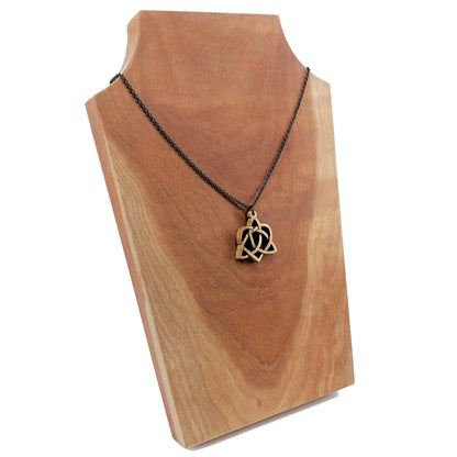 Wooden necklace pendant created in the style of a Celtic weave pattern that is a trinity and heart combined. The symbol represents adoption. Hanging from a silver stainless steel chain against a cherry wood display.