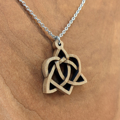Wooden necklace pendant created in the style of a Celtic weave pattern that is a trinity and heart combined. The symbol represents adoption. Hanging from a silver stainless steel chain against a cherry wood background.