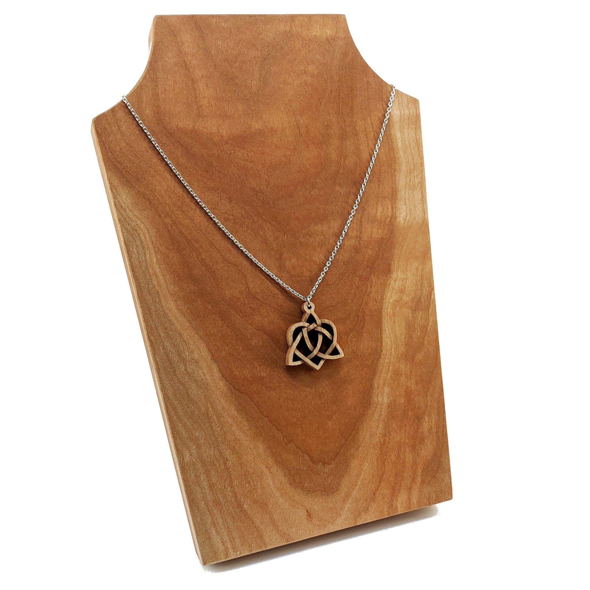 Wooden necklace pendant created in the style of a Celtic weave pattern that is a trinity and heart combined. The symbol represents adoption. Hanging from a silver stainless steel chain against a cherry wood display.