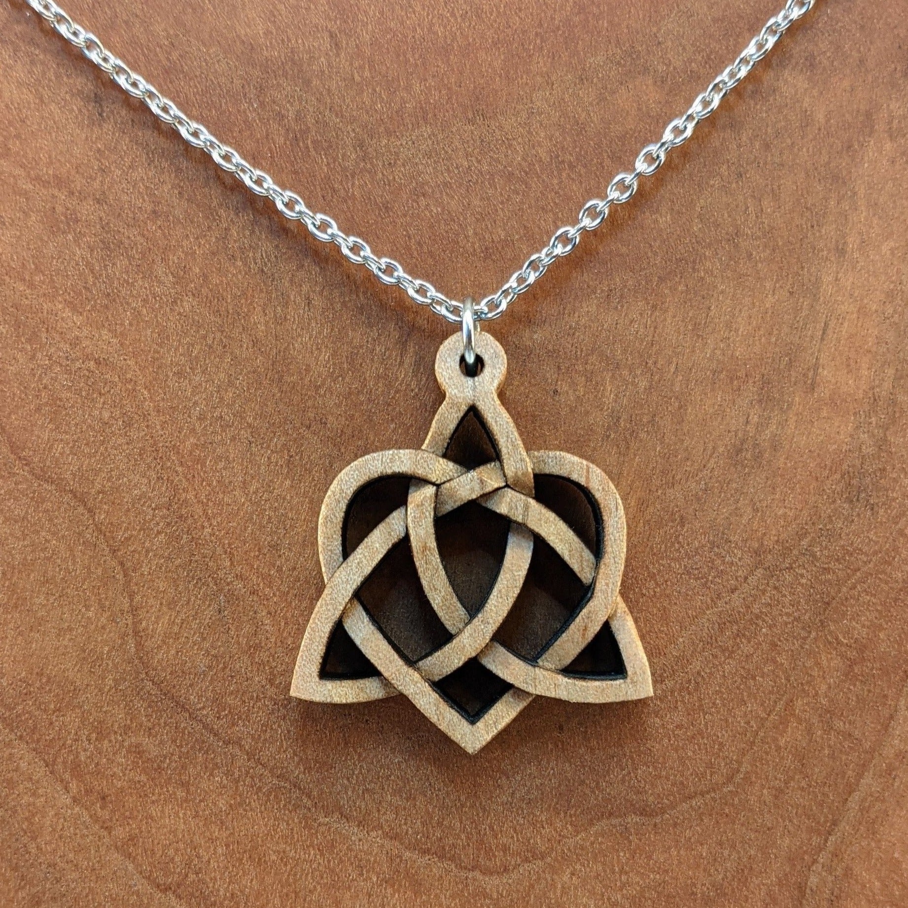 Wooden necklace pendant created in the style of a Celtic weave pattern that is a trinity and heart combined. The symbol represents adoption. Hanging from a silver stainless steel chain against a cherry wood background.