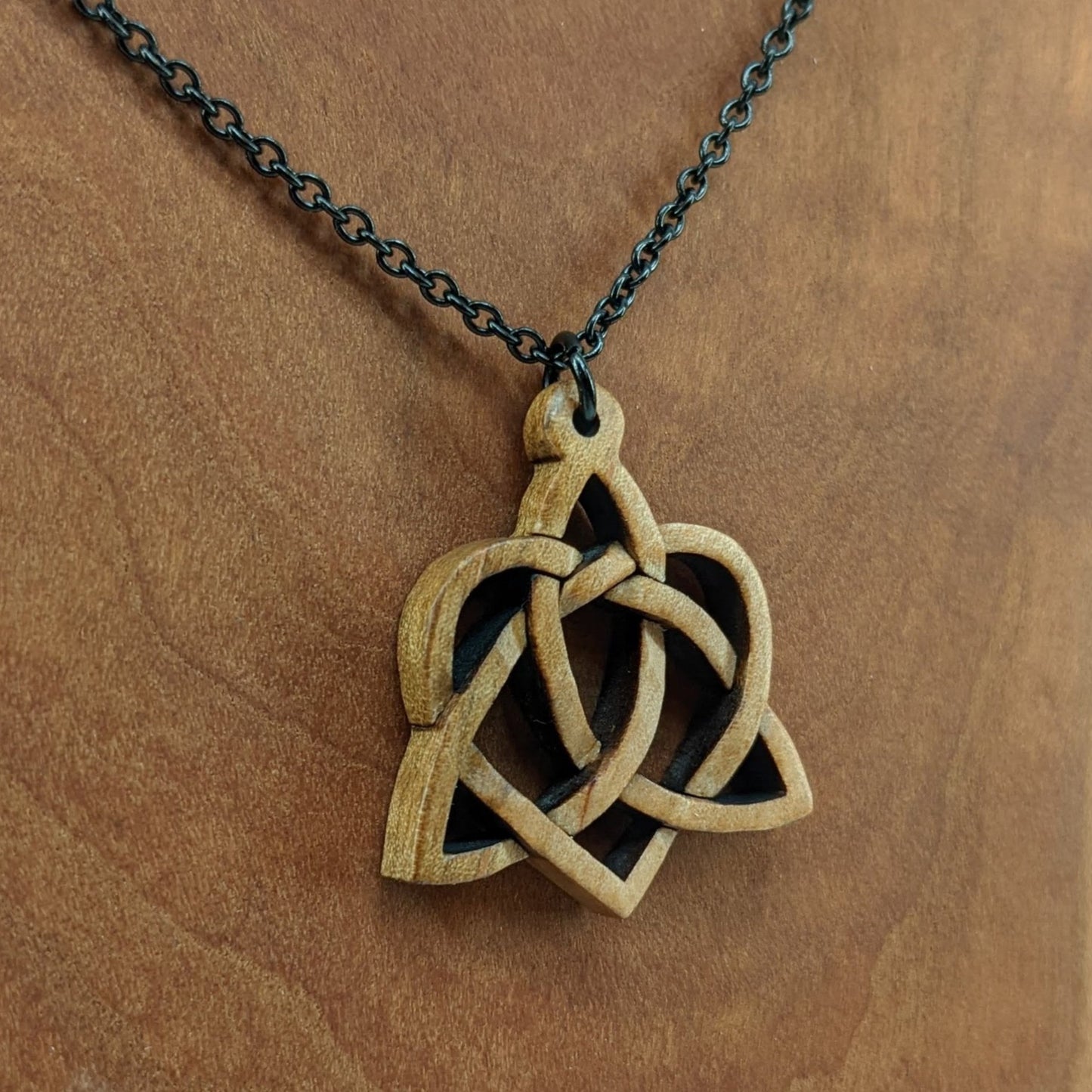 Wooden necklace pendant created in the style of a Celtic weave pattern that is a trinity and heart combined. The symbol represents adoption. Hanging from a black stainless steel chain against a cherry wood background.