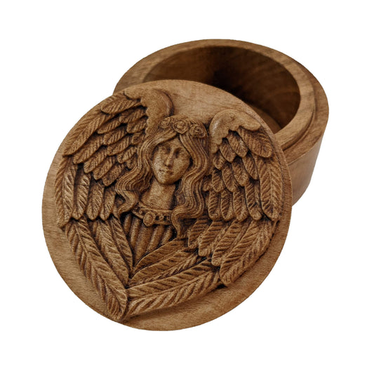 Round wooden box 3D carved with a intricately winged angel wearing a jeweled necklace and flowers in her hair. Her wings spread from behind her and wrap around her shoulders. Made from hard maple wood with a brown stain against a white background.