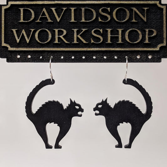 Pair of wooden earrings with silver stainless steel hooks hanging on a display. They are silhouettes of tensed, hissing black cats. Made from birch wood hanging from a model Davidson Workshop sign against a white background.