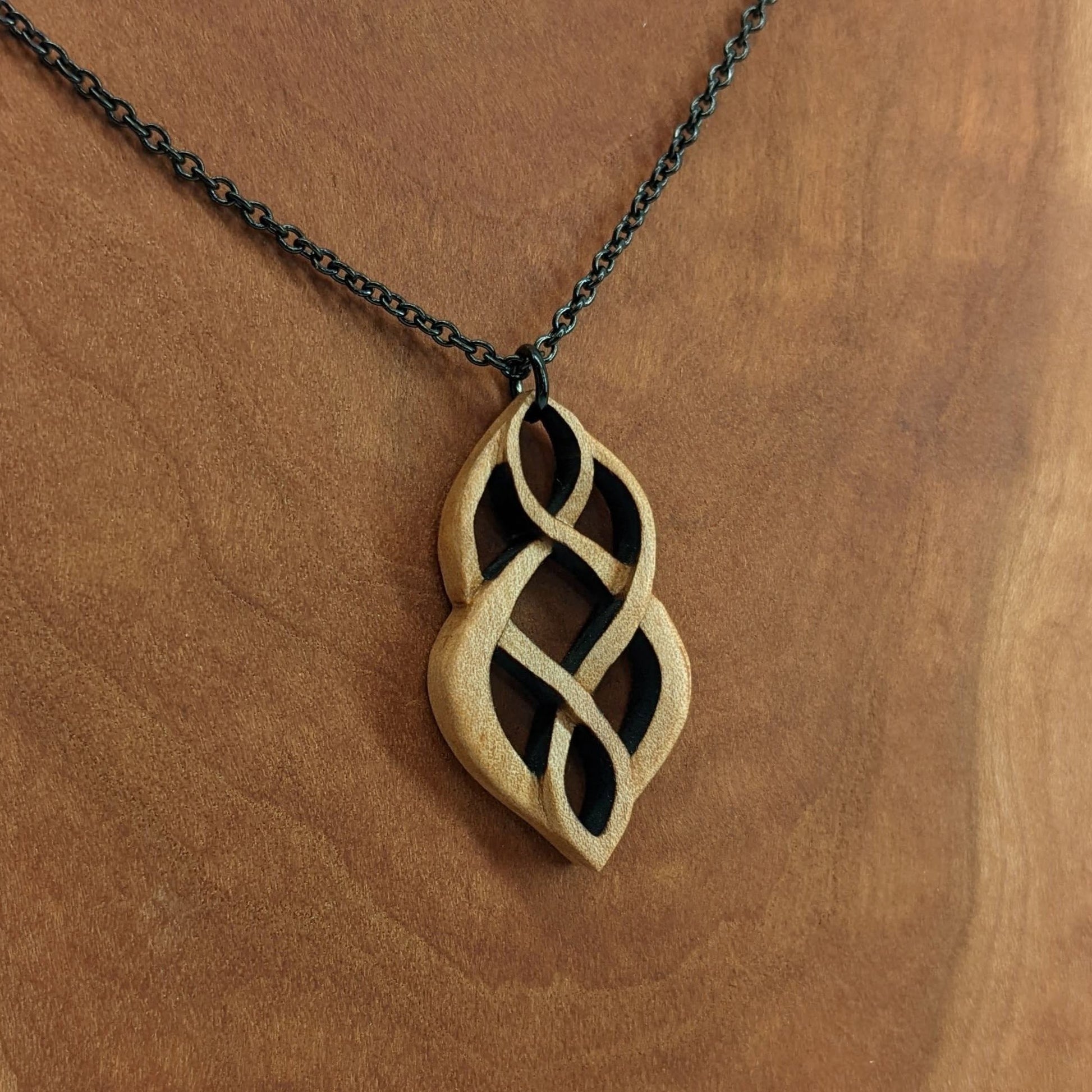 Wooden necklace pendant carved so that curving shapes weave into each other. Made from hard maple wood and hanging from a black chain against a cherry wood background.