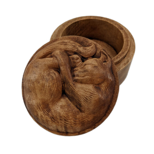 Round wooden box 3D carved with a short haired cat with textured fur curled up into a ball sleeping. It has a peaceful rested expression on its face and legs all crossed together. Made from hard maple wood stained brown against a white background.