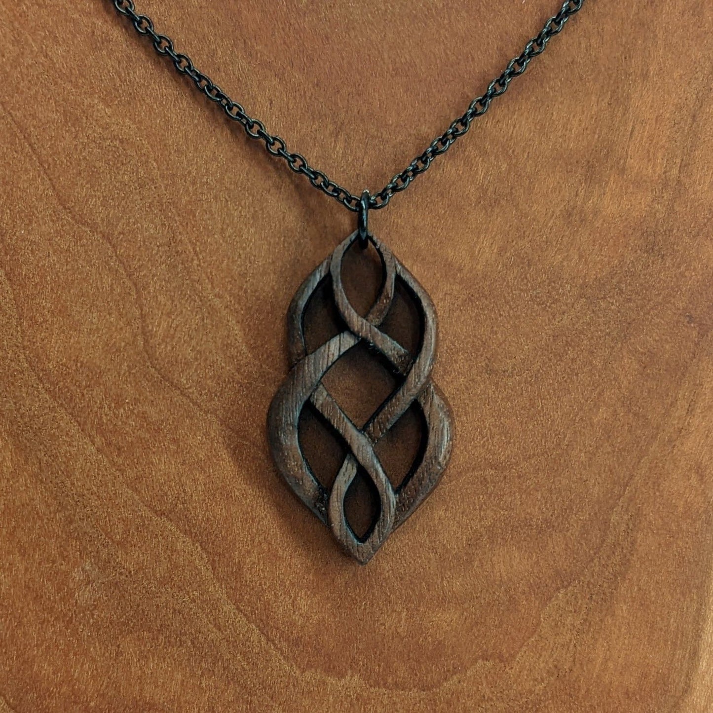 Wooden necklace pendant carved so that curving shapes weave into each other. Made from dark walnut wood and hanging from a black chain against a cherry wood walnut background.