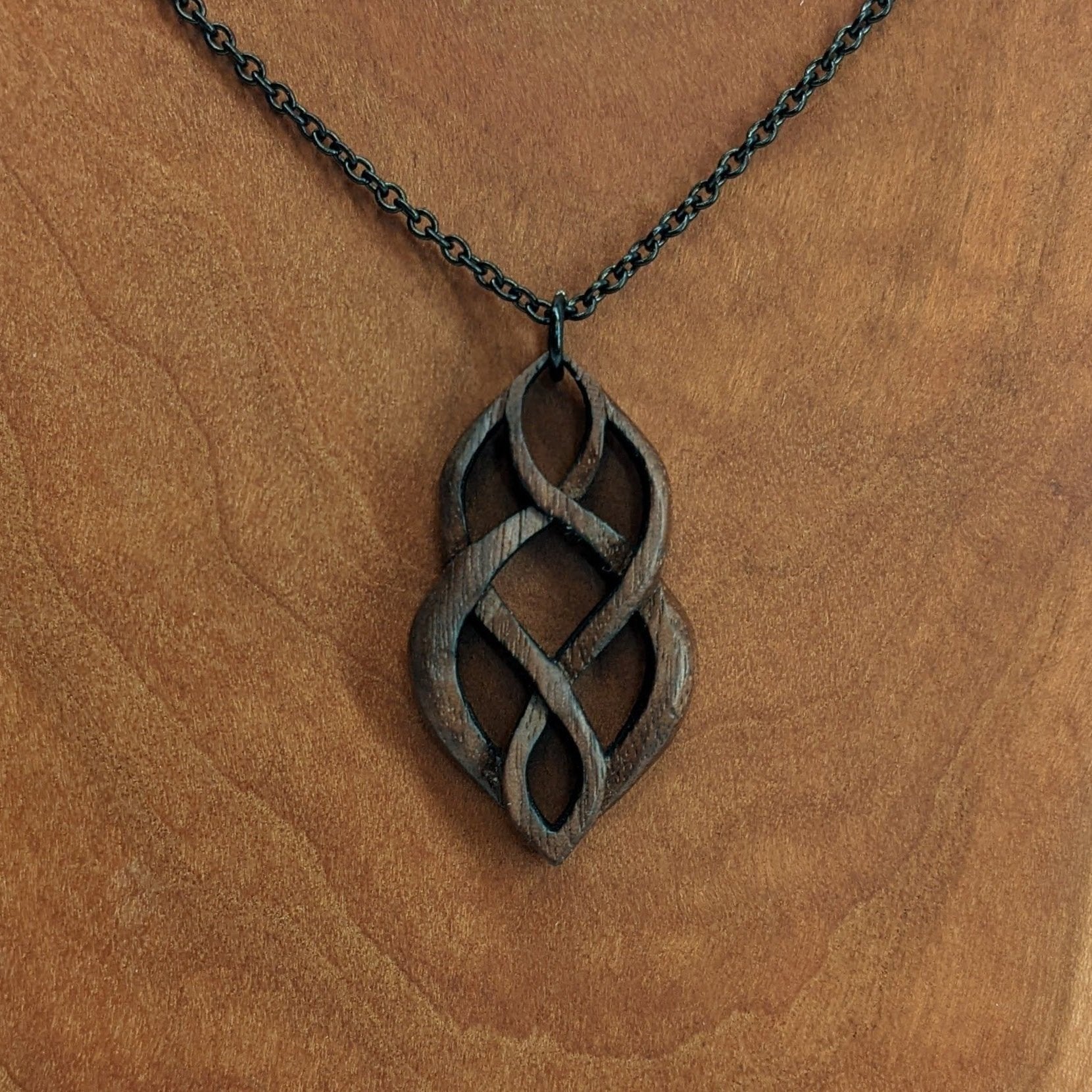 Wooden necklace pendant carved so that curving shapes weave into each other. Made from dark walnut wood and hanging from a black chain against a cherry wood walnut background.