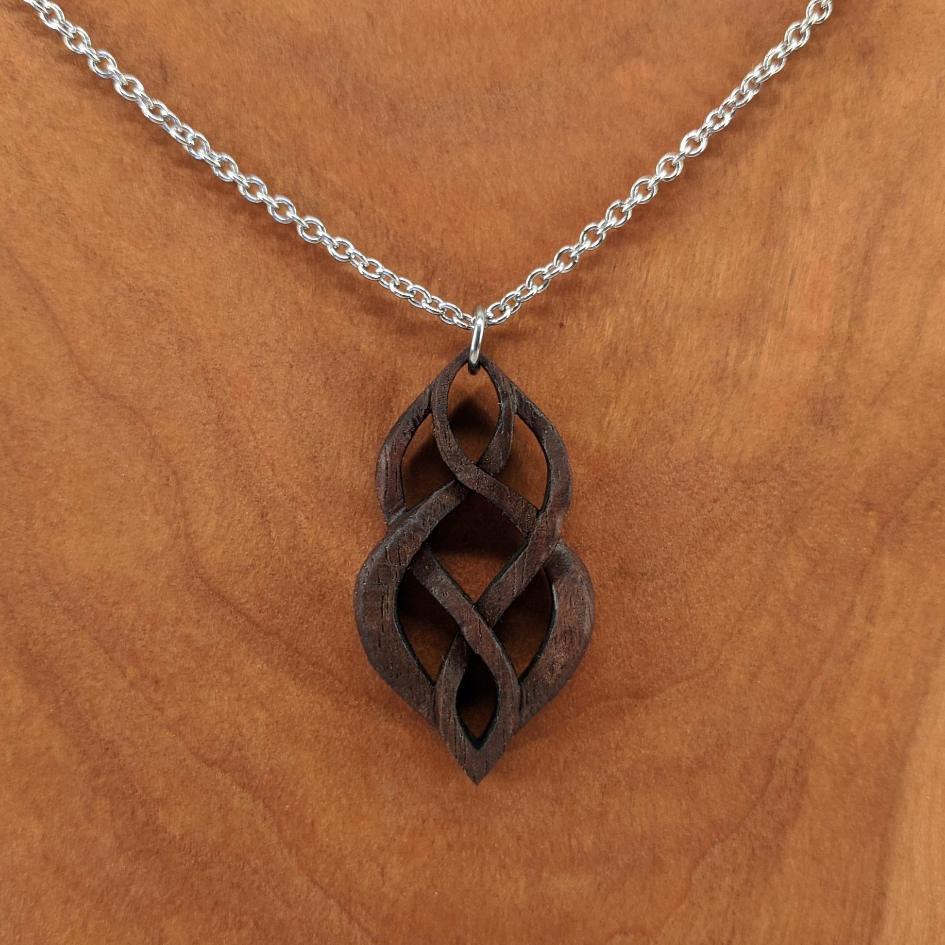 Wooden necklace pendant carved so that curving shapes weave into each other. Made from dark walnut wood and hanging from a silver chain against a cherry wood walnut background.