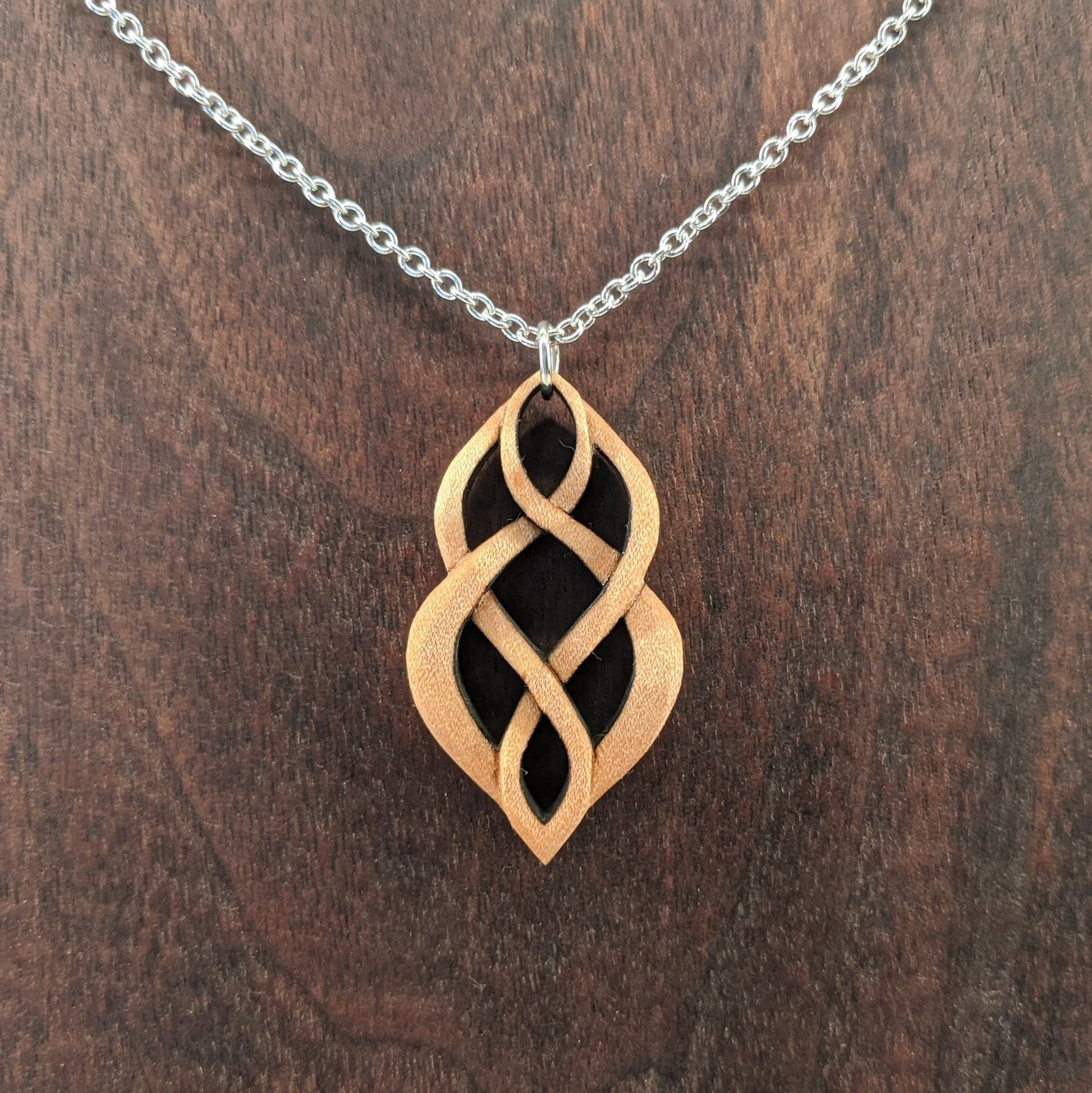 Wooden necklace pendant carved so that curving shapes weave into each other. Made from hard maple wood and hanging from a silver chain against a dark walnut background.