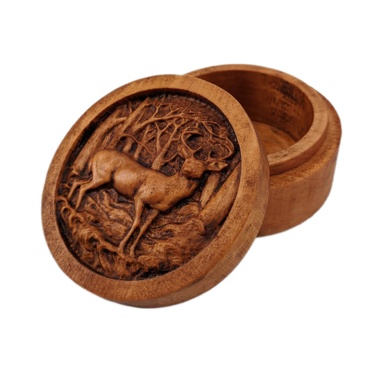 Round wooden box 3D carved with a buck deer standing in a forest. It stands in small grassy knolls in front of trees that have thin branches sticking out in every direction. Made from hard maple wood with a brown stain against a white background.