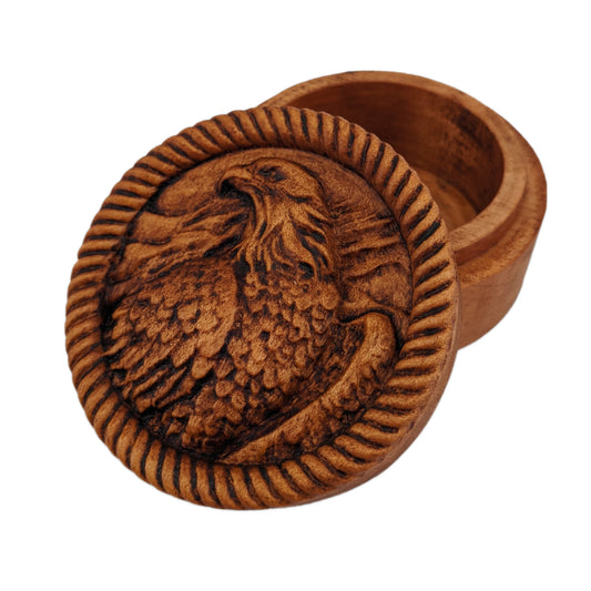Round wooden box 3D carved with a bald eagle over a wavy background surrounded by a rope border. It has flowing feathers around its head and layered ones on the chest and wings. Made from hard maple wood with a brown stain against a white background.