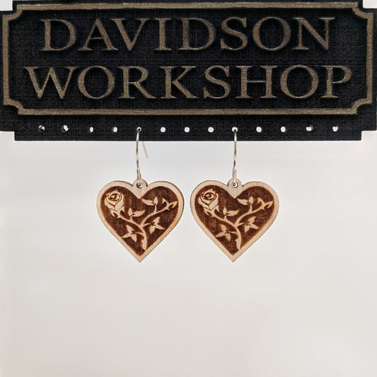 Pair of wooden earrings with silver stainless steel hooks. They are two toned light and dark brown rose and vines inside of a heart shape. Made from birch wood hanging from a model Davidson Workshop sign against a white background. (Close up View)