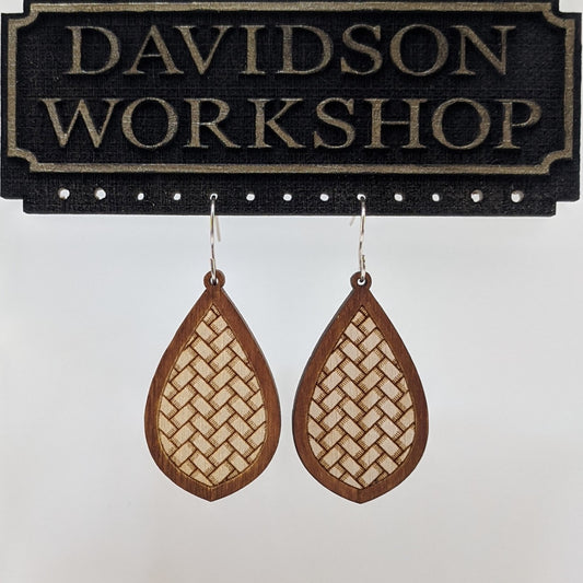 Pair of wooden earrings with silver stainless steel hooks. They are brown almond shaped with an engraved herringbone weave pattern within. Made from birch wood hanging from a model Davidson Workshop sign against a white background. (Close up View)
