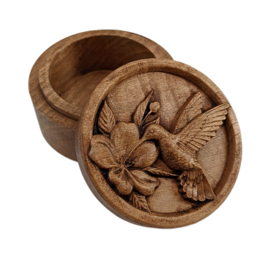 Round wooden box 3D carved with a recessed hummingbird hovering and drinking nectar from a flower. It is facing to the left with its delicate wings aflutter as it leans in to drink. Made from hard maple wood stained brown against a white background.