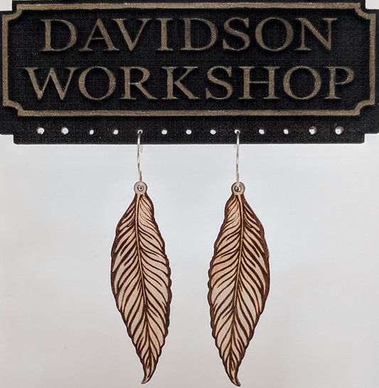 Pair of wooden earrings with silver stainless steel hooks. They are pale natural finished, long and slim realistic engraved feathers. Made from birch wood hanging from a model Davidson Workshop sign against a white background. (Close up View)