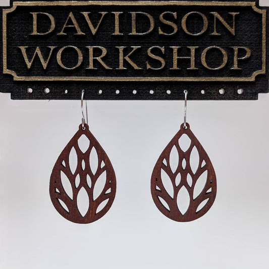 Pair of wooden earrings with silver stainless steel hooks. They are brown teardrop shaped. With multiple eye and almond shaped cutouts within. Made from birch wood hanging from a model Davidson Workshop sign against a white background (Close up View)