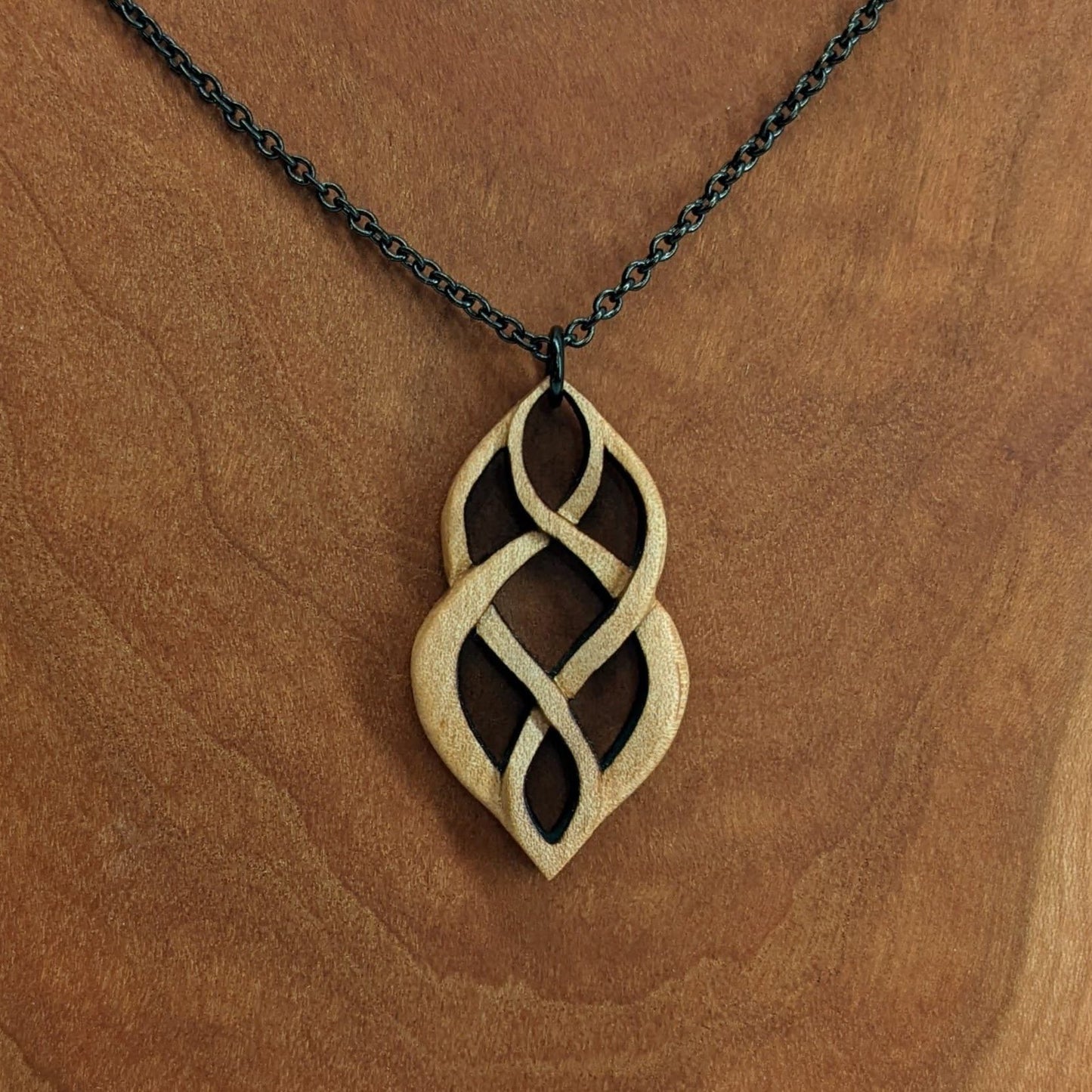 Wooden necklace pendant carved so that curving shapes weave into each other. Made from hard maple wood and hanging from a black chain against a cherry wood background.