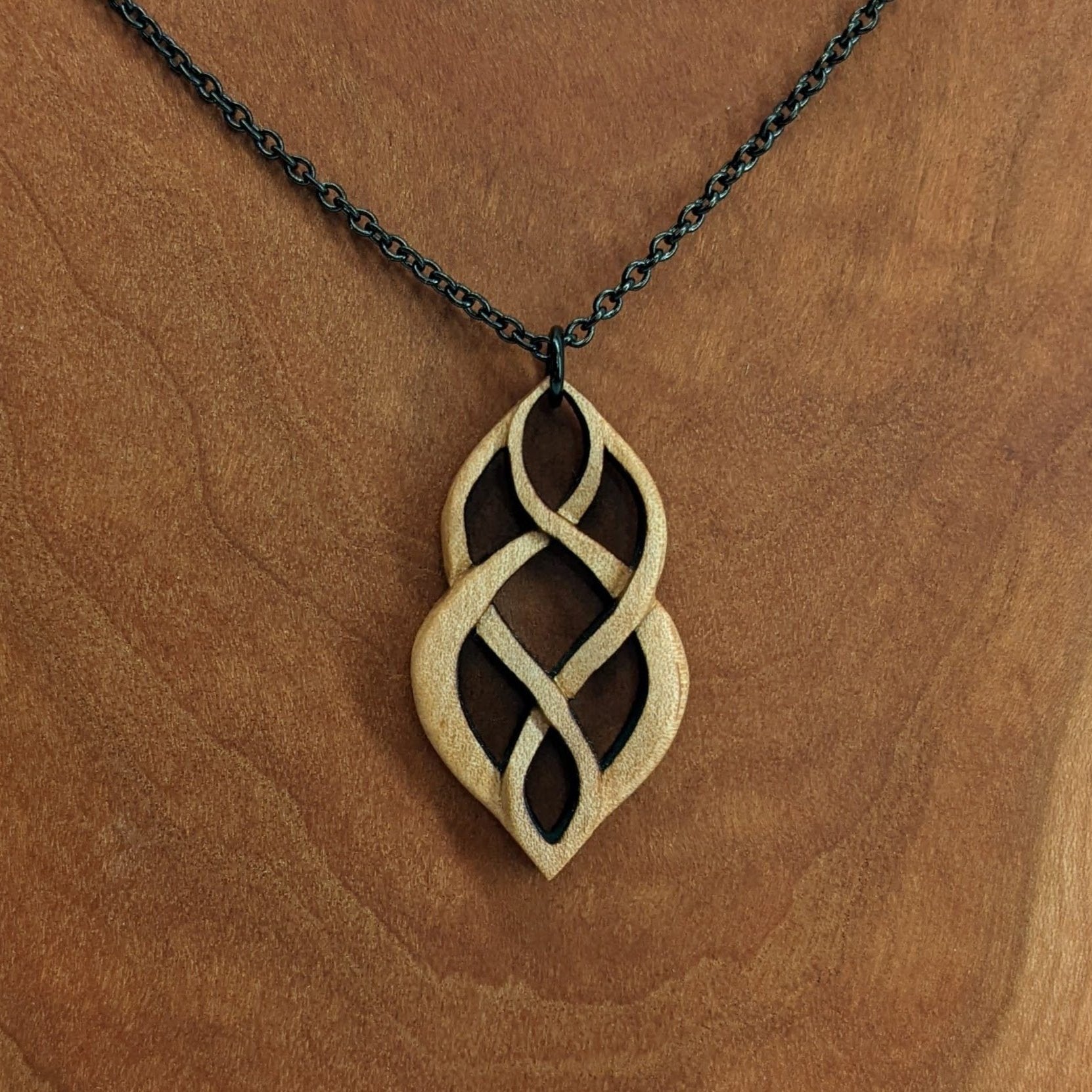 Wooden necklace pendant carved so that curving shapes weave into each other. Made from hard maple wood and hanging from a black chain against a cherry wood background.