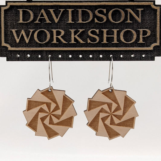 Pair of wooden earrings with silver stainless steel hooks. They are light natural finish pinwheels with engraved overlapping blades. Made from birch wood hanging from a model Davidson Workshop sign against a white background. (Close up View)