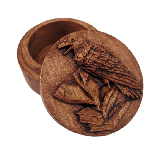 Round wooden box 3D carved with a raven standing three books with a quill at its feet, the top book open and pages  turning. The raven has an open beak and intricate layered feathers Made from hard maple wood stained brown against a white background.