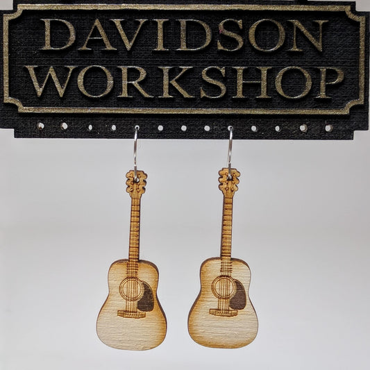 Pair of wooden earrings with silver stainless steel hooks. They are light natural finish guitars with engraved strings and other details. Made from birch wood hanging from a model Davidson Workshop sign against a white background. (Close up View)