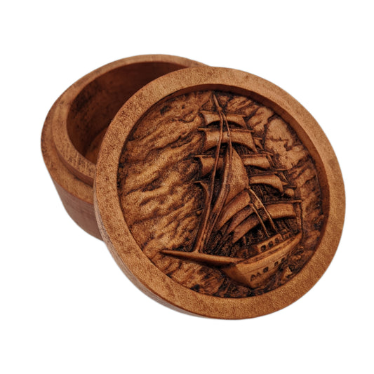 Round wooden box 3D carved with a large sailboat on the sea. It has multiple sails all pulled taught with ropes hanging all around. The background is a cloudy sky and rolling waves. Made from hard maple wood stained brown against a white background.