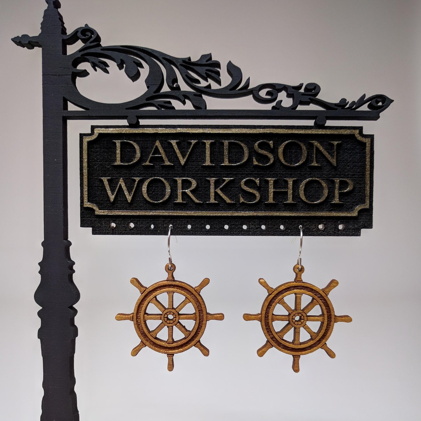 Pair of wooden earrings with silver stainless steel hooks. They are brown pirate ship wheels with 8 handles and a spoked center. Made from birch wood hanging from a model Davidson Workshop sign against a white background.