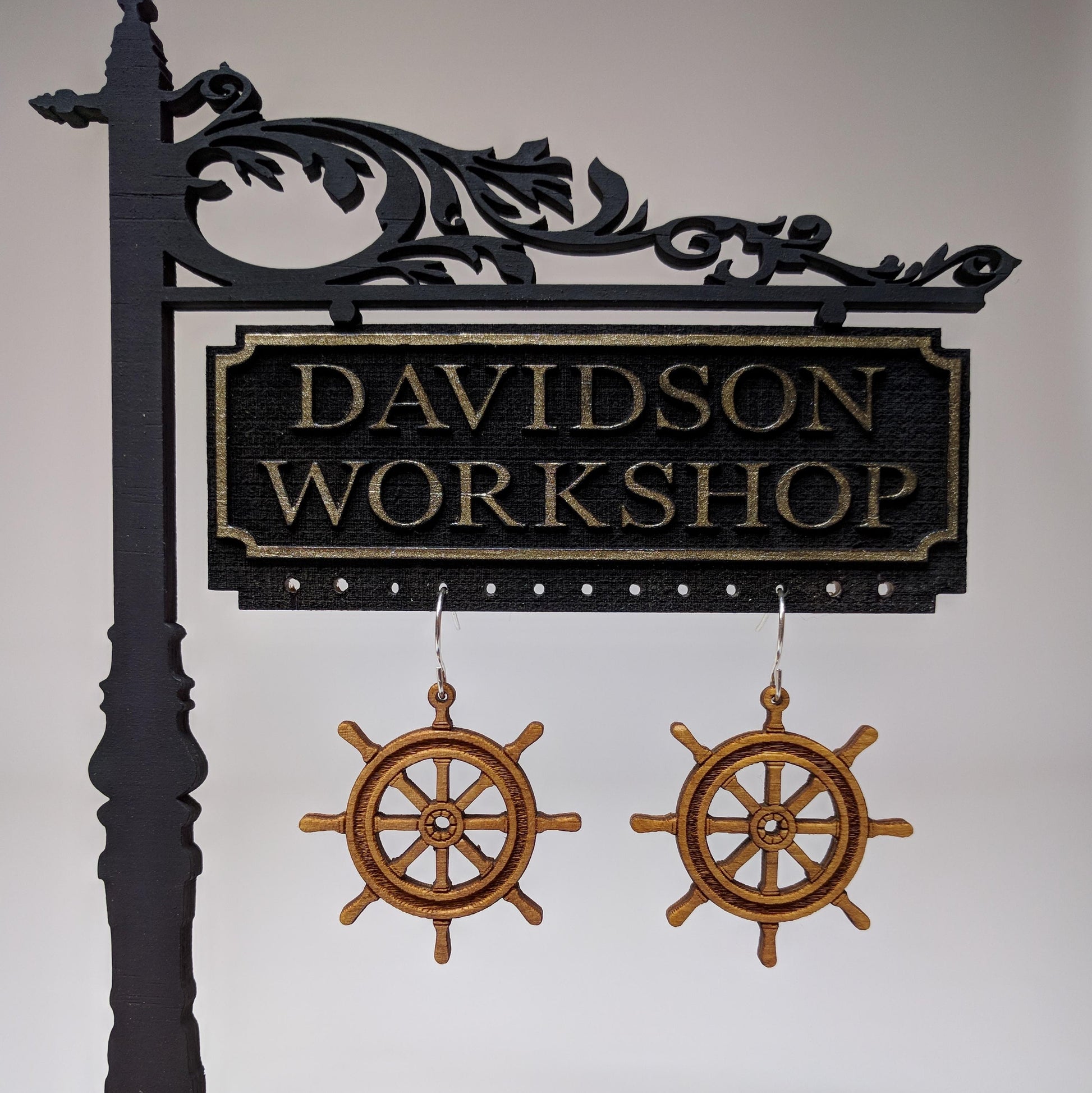 Pair of wooden earrings with silver stainless steel hooks. They are brown pirate ship wheels with 8 handles and a spoked center. Made from birch wood hanging from a model Davidson Workshop sign against a white background.