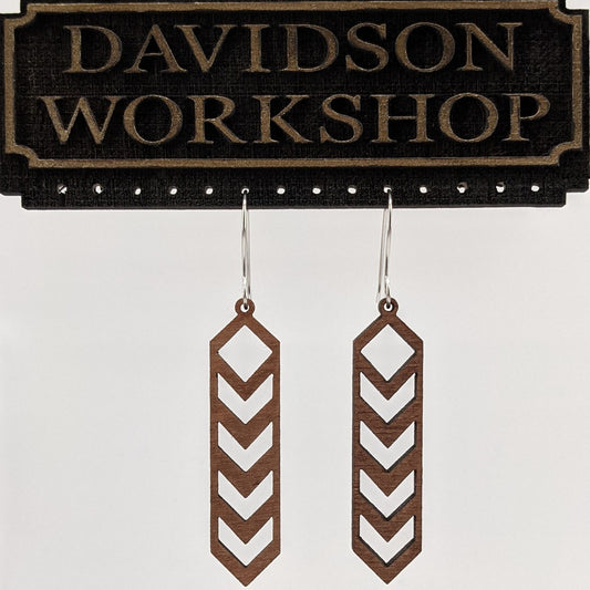 Pair of wooden earrings with silver stainless steel hooks. They are brown slim, elongated diamond shapes with downward chevron cutouts within. Made from birch wood hanging from a model Davidson Workshop sign against a white background.(Close up View)