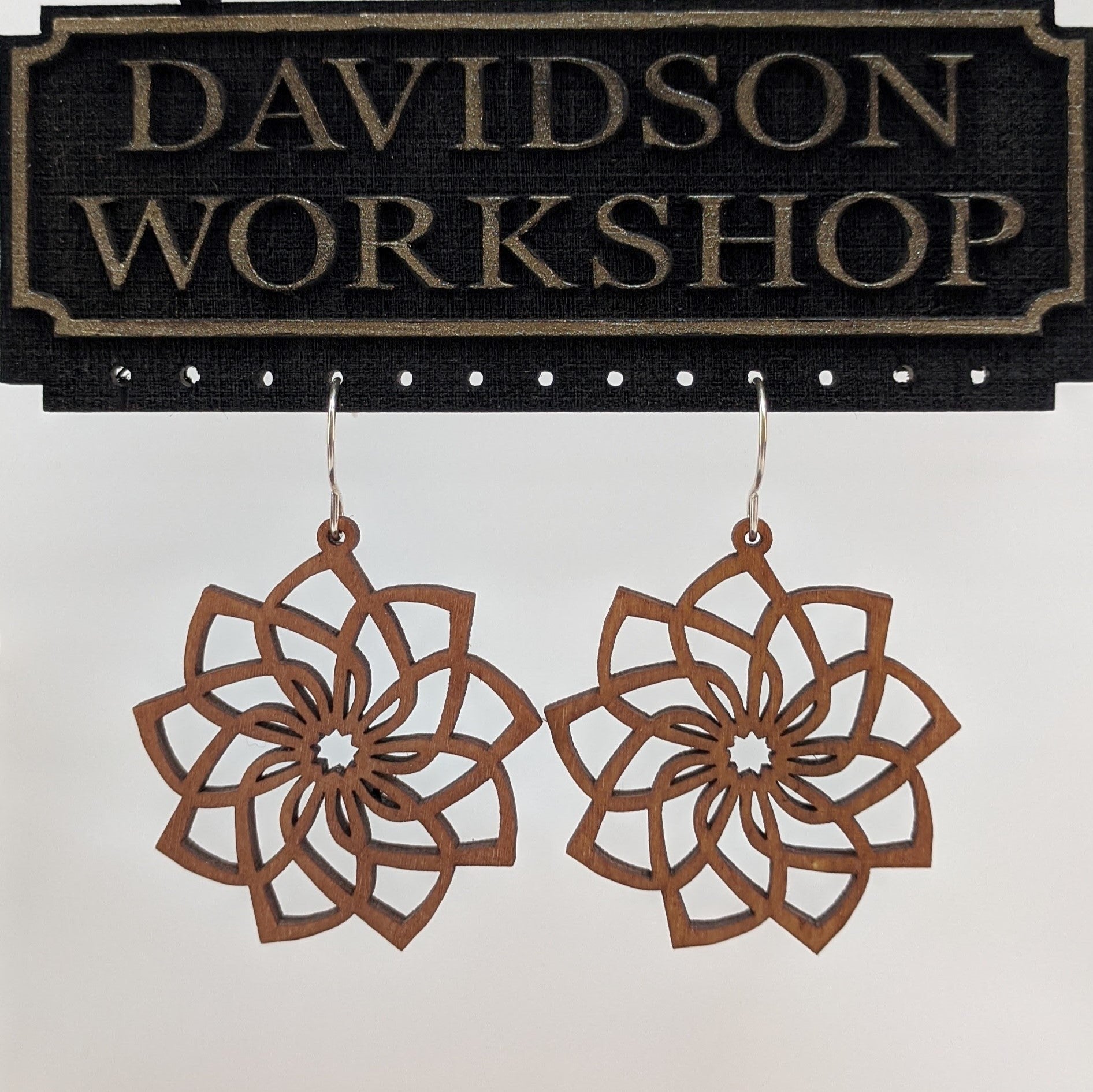 Pair of wooden earrings with silver stainless steel hooks. They are brown cutouts of flowers with spiraling, overlapping petals. Made from birch wood hanging from a model Davidson Workshop sign against a white background. (Close up View)