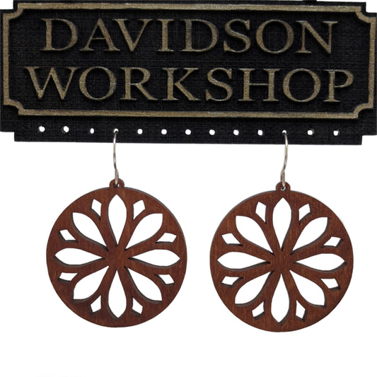 Pair of wooden earrings with silver stainless steel hooks. They are brown round with symmetrical petal shaped cutouts within. Made from birch wood hanging from a model Davidson Workshop sign against a white background. (Close up View)