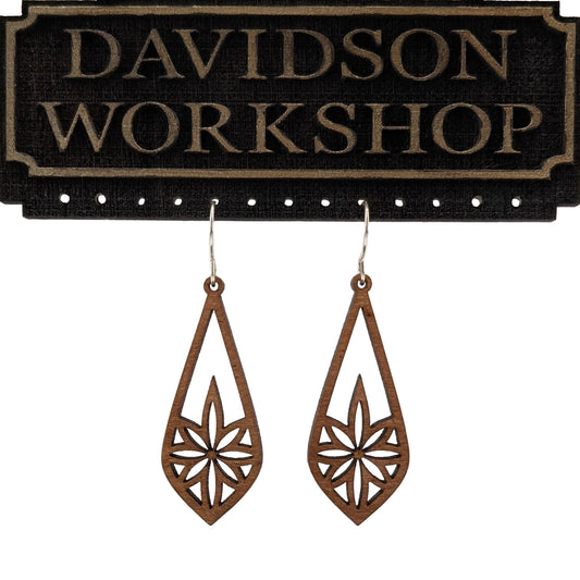 Pair of wooden earrings with silver stainless steel hooks. They are long brown teardrops with an 8 pointed star with sharp points within. Made from birch wood hanging from a model Davidson Workshop sign against a white background. (Close up View)