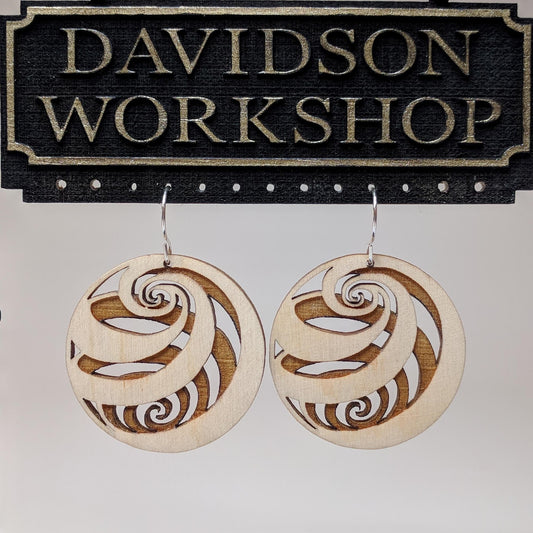 Pair of wooden earrings with silver stainless steel hooks. They are light finish balls with spirals engraved creating a 3D spherical illusion. Made from birch wood hanging from a model Davidson Workshop sign against a white background.(Close up View)