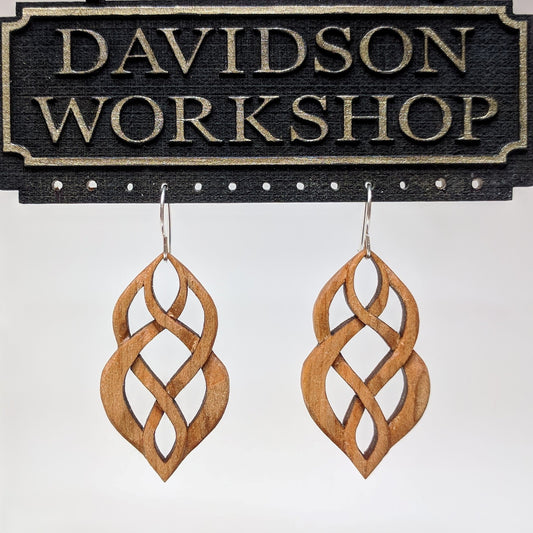 Pair of wooden earrings with silver stainless steel hooks. They are Celtic style, carved in a woven pattern to form an hourglass shape. Made from hard maple hanging from a model Davidson Workshop sign against a white background. (Close up View)