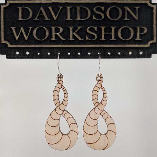 Pair of wooden earrings with silver stainless steel hooks. They are light natural finish scaled infinity symbols twisting over themselves.  Made from birch wood hanging from a model Davidson Workshop sign against a white background. (Close up View)