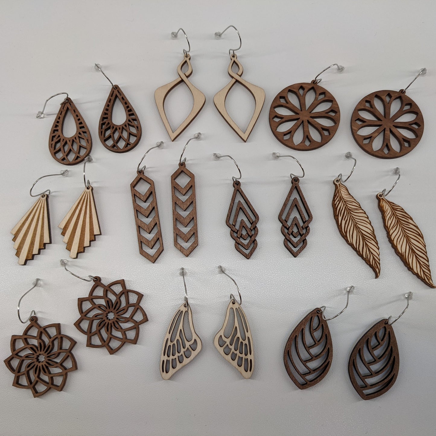 10 pairs of wooden earrings with silver stainless steel hooks. There are multiple designs in both light natural finish and dark brown, styles from nature inspired to ornate geometric patterns. Made from birch wood, laying against a white background.