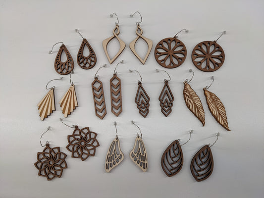 10 pairs of wooden earrings with silver stainless steel hooks. There are multiple designs in both light natural finish and dark brown, styles from nature inspired to ornate geometric patterns. Made from birch wood, laying against a white background. (Close up view)