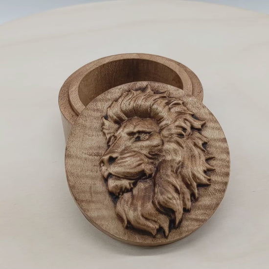 Round wooden box 3D carved with a side profile of a lions face. It has a serious look in its brow with a closed mouth facing to the left with a large bushy mane surrounding its face. Made from hard maple wood stained brown against a white background rotating on a table.