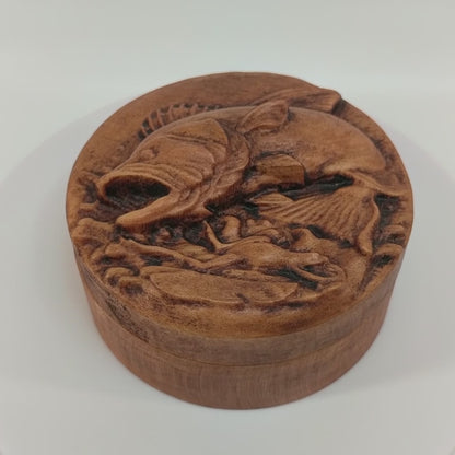 Round wooden box 3D carved with a large bass fish jumping out of the water chasing after a small frog that has narrowly avoided being caught. Both are overtop a watery splash. Made from hard maple wood with a brown stain against a white background rotating on a table.