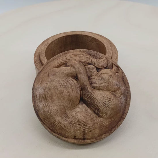 Round wooden box 3D carved with a short haired cat with textured fur curled up into a ball sleeping. It has a peaceful rested expression on its face and legs all crossed together. Made from hard maple wood stained brown against a white background rotating on a table.