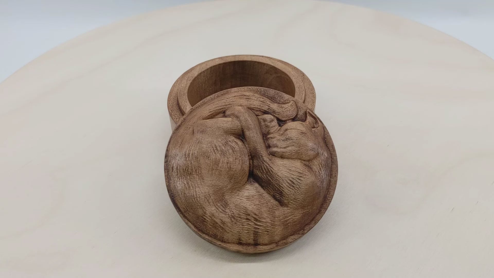 Round wooden box 3D carved with a short haired cat with textured fur curled up into a ball sleeping. It has a peaceful rested expression on its face and legs all crossed together. Made from hard maple wood stained brown against a white background rotating on a table.