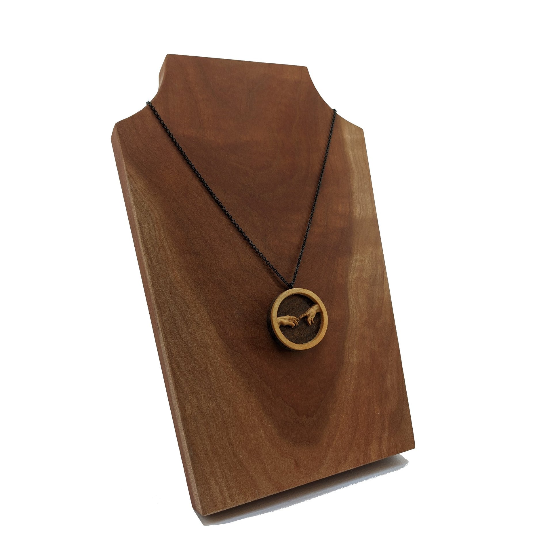 Round wooden necklace pendant carved in the shape of two hands reaching out to touch each other. The hands are carved from light maple wood overlayed on a dark walnut base. Hanging from a black stainless steel chain against a cherry wood display.