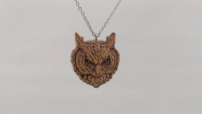 Wooden necklace pendant carved in the shaped of an owls head. Made from hard maple wood. Hanging and rotating from a silver stainless steel chain against a white background.